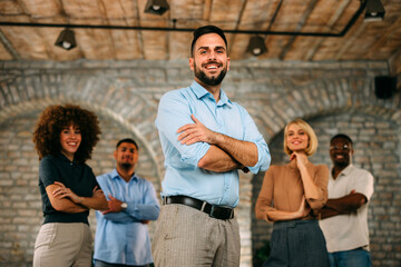 Confident and smiling male leader, crossed arms, standing in front of his diverse team of young professionals in a modern, rustic office environment.