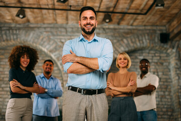 Confident male leader with crossed arms smiling in front of his diverse, multi-ethnic business team in a modern office