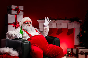 Santa relaxing in red Christmas lounge with beer in hand amid gifts and stockings by a warm...