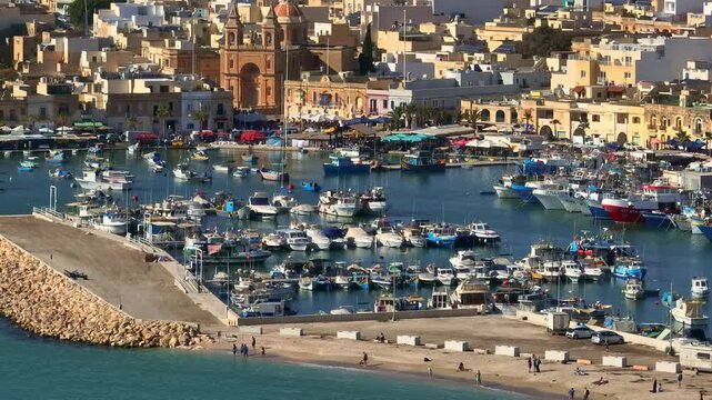 Malta, Marsaxlokk Harbour: Colorful boats fill the picturesque coastline harbor bay creating vibrant scene against the backdrop of charming town in sunny day. Travel destination. Aerial drone flight