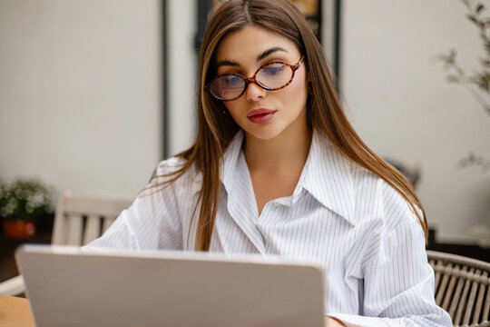 Close-up of young woman in glasses working on laptop at outdoor cafe. Focused freelancer using computer for business or studying, modern technology concept. - Powered by Adobe