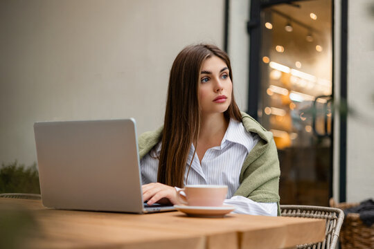 Thoughtful young woman working on laptop at outdoor cafe, wearing light green sweater. Freelancer taking a break, creative thinking and remote work lifestyle concept.