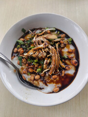 A white bowl of Indonesian chicken porridge (bubur ayam) topped with shredded chicken, peanuts, and fried shallots on a wooden table.