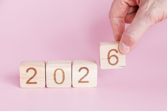 Hand is lifting wooden block with number six to replace five in sequence indicating transition from 2025 to 2026 on soft pink backdrop.