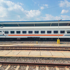 A modern white passenger train car with orange stripes parked at a sunny outdoor railway station.