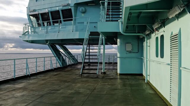 empty ship deck under cloudy sky, metal staircase and railings leading to elevated superstructure, wet worn surface with rust stains, distant choppy ocean.