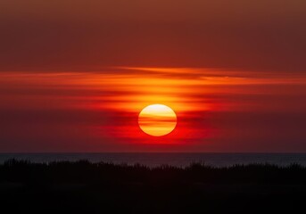 Fototapeta premium Intense red and orange sunset through thick atmospheric haze creating a dramatic, warped, and misshapen solar disk near the horizon line ,wildfire smoke ,twilight ,dusk