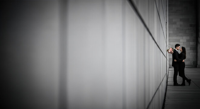 Young, elegant couple sharing an intimate moment, embracing tightly against a modern, dramatic gray architectural wall with ample negative copy space.