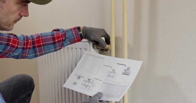 Gloved technician holds schema by wall near radiator pipes. Plumber verifies connection route selecting parts and preparing assembly sequence during maintenance work