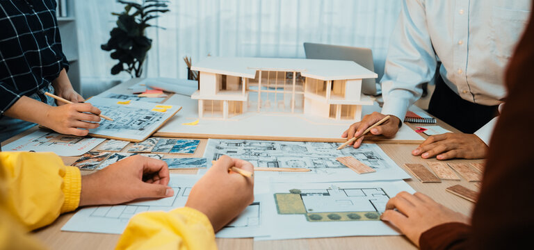 A group of architects and designers engage in a collaborative design session, surrounded by architectural plans and a detailed model. The bright office fosters creativity. SACTR - Powered by Adobe