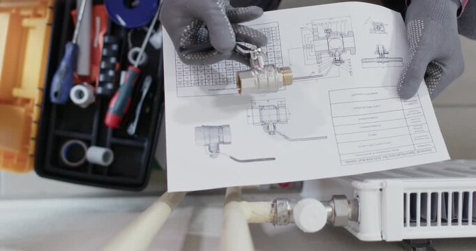 Male technician hands hold ball valve above schematic sheet reviewing layout. Plumber matches connector size and checks part number marking install steps