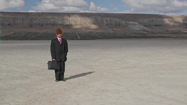 A curly-haired teenager with a sad face and a mature business suit stands on dry ground with a briefcase against the backdrop of a hill with a vineyard.