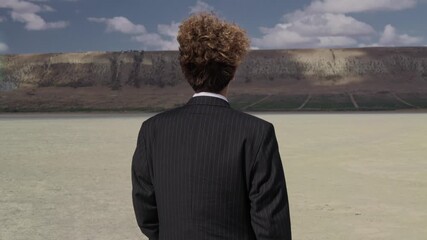A teenager in a business suit walks against a blue sky, curls fluttering in the wind. View from behind