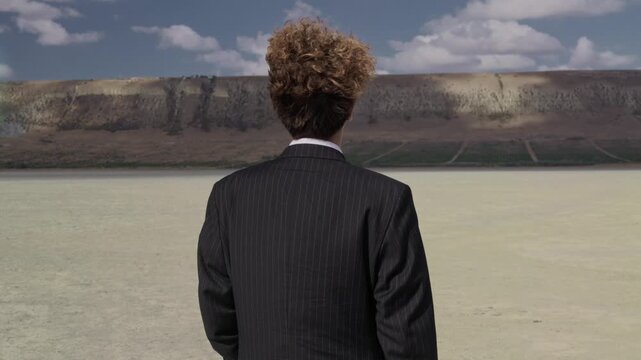 A teenager in a business suit walks against a blue sky, curls fluttering in the wind. View from behind