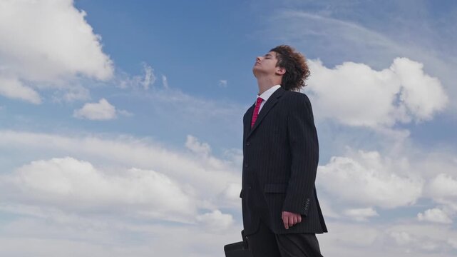 Portrait of a handsome teenager in a business suit against a blue sky, his face upturned and his curls fluttering in the wind. The camera pans.