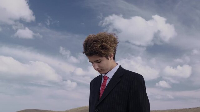 Portrait of a handsome, serious teenager in a business suit against a blue sky, his head down and curls fluttering in the wind
