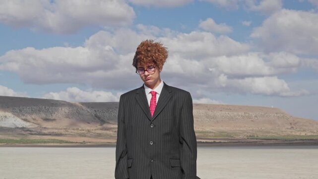A curly-haired teenager with a sad face and a mature business suit stands on dry ground with a briefcase against the backdrop of a hill with a vineyard. The camera zooms out.