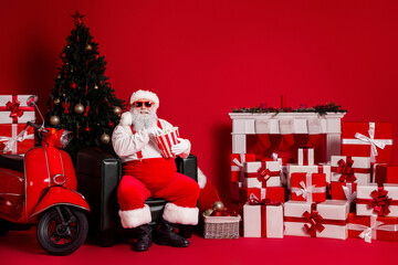 Santa sits amid gifts and a Christmas tree beside a red backdrop with a scooter and fireplace scene...