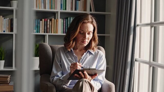 Sunlit library armchair writer using tablet, bookshelf backdrop, tall window casting soft natural light, pen interaction with touchscreen, focused expression, stack of books and potted plant, calm
