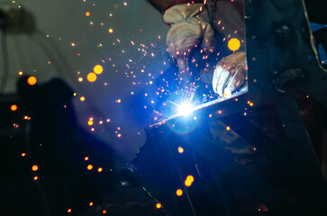 Welder works welding steel parts at factory in protective gloves. Smoke, sparks, blue light.