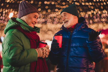 Warm moment between a senior couple enjoying coffee cups under twinkling holiday lights in a lively city market during winter festive