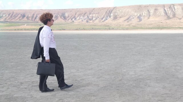 A teenager in a business suit walks against a backdrop of hills and a blue sky, his curls fluttering in the wind. He stops and expresses displeasure