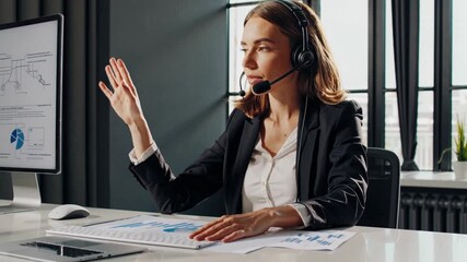 white businesswoman on video call wearing headset and blazer seated at modern desk with laptop and paperwork charts visible on monitor gesturing to participants listening attentively and raising hand - Powered by Adobe
