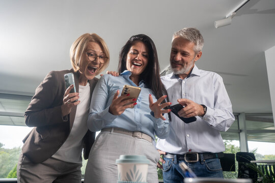 Excited diverse business team standing together looking at smartphone celebrating success