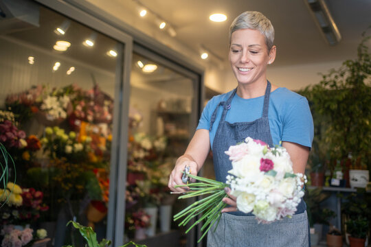 Smiling female florist cutting and arranging bouquet of fresh flowers for customers