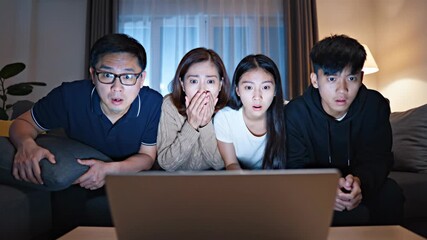 Asian family watching a scary movie or shocking content on a laptop at home during the night, expressing fear, shock, and suspense while sitting on the sofa