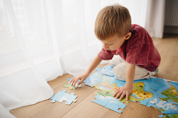 A young child focuses on completing a colorful puzzle featuring a world map. Surrounding pieces are scattered on a wooden floor, creating a playful learning environment. top view