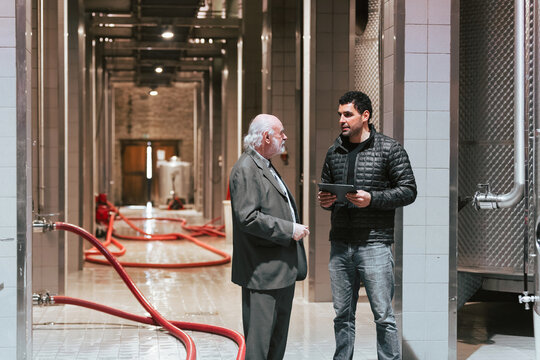 Two men converse about wine production in a winery surrounded by tanks and hoses.