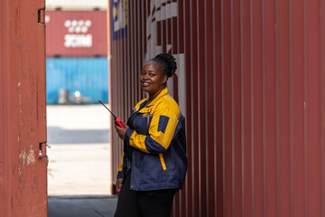 Female logistics worker standing between cargo containers, holding walkie talkie and smiling at shipping yard.