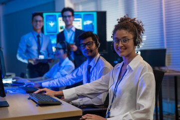 Diverse team of professionals customer service agent wearing headsets and monitoring computer working in dark control room. Female support and assistance call center at night in modern office.