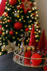 Beautiful Christmas composition with red decorative trees, a golden reindeer figurine, and shiny gold ornaments on a golden tray. In the background — a glowing Christmas tree with warm fairy lights. 