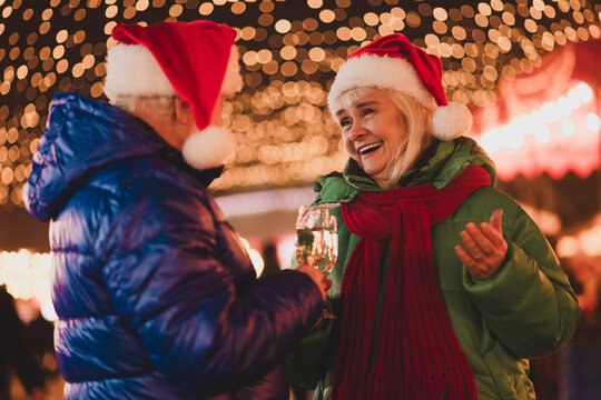 Festive Santa couple sharing a toast outdoors under twinkling Christmas lights during a winter market scene full of warmth smiles and holiday joy - Powered by Adobe