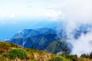 Majestic mountain scenery of Pico do Arieiro in Madeira, Portugal, rugged peaks extending towards the Atlantic Ocean. Lush foreground vegetation and dramatic clouds rolling over the peaks of island