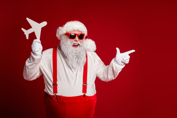 Cheerful Santa with red suspenders and hat holds plane cutout against red background for Christmas celebration festive marketing image