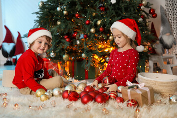 Cute children in Santa hats with ornaments near Christmas tree at home