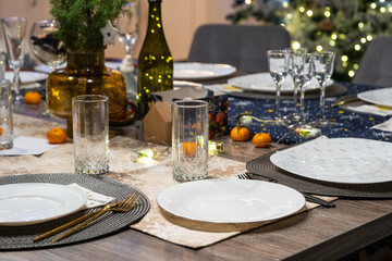 A festive table set for guests for Christmas and New Year, a Christmas tree in the living room, plates, bottles, glasses, potted plants, fairy lights