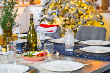 A festive table set for guests for Christmas and New Year, a Christmas tree in the living room, plates, bottles, glasses, potted plants, fairy lights