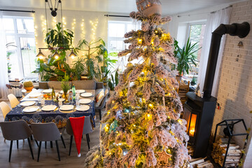 Festive interior of house is decorated for Christmas and New Year in loft style with black stove, fireplace, Christmas tree. Warm studio room with set table, burning wood, cozy and heating of home