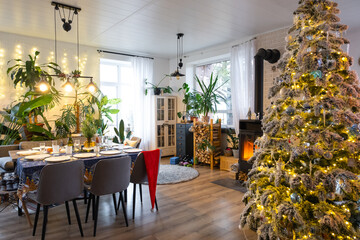 Festive interior of house is decorated for Christmas and New Year in loft style with black stove, fireplace, Christmas tree. Warm studio room with set table, burning wood, cozy and heating of home
