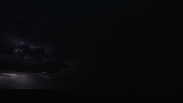 Dark night sky illuminated by distant lightning flickering through storm clouds 