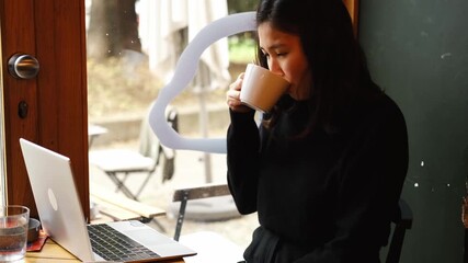 Young asian woman drinks cappuccino while working on her laptop in a cozy cafe by the window, enjoying a peaceful morning atmosphere. Horizontal 4k footage - Powered by Adobe