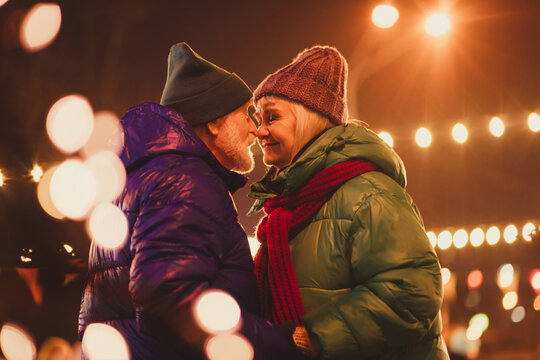 Couple of grandparents share a warm Christmas moment under twinkling lights in a cozy winter city scene at a festive outdoor fair - Powered by Adobe