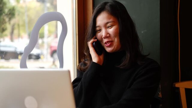 Asian woman smiles while talking on the phone and looking at her laptop in a cozy cafe by the window. Morning work and communication scene. Horizontal 4k footage