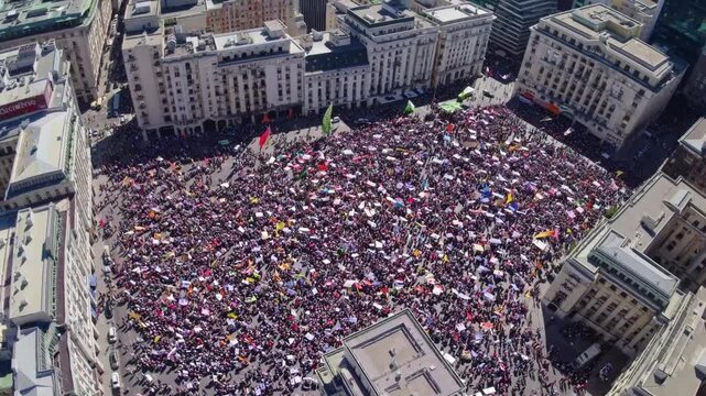 Mass aerial crowd fills city plaza with colorful signs and flags, densely packed protesters surrounded by historic buildings, daytime sunlight, slow panoramic sweep revealing scale, peaceful unity