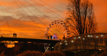 People silhouettes walking on bridge during golden hour sunset with Ferris wheel and Christmas lights in background