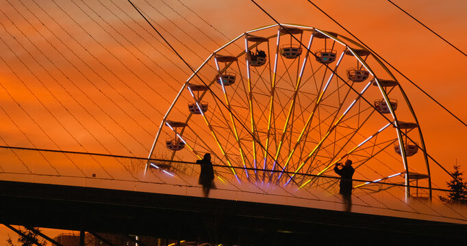 Silhouetted tourists admire illuminated Ferris wheel from pedestrian bridge during golden hour